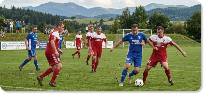 Futbalový zápas dvoch tímov v modrých a červených dresoch počas regionálneho turnaja v prírodnom prostredí s pohorím v pozadí.